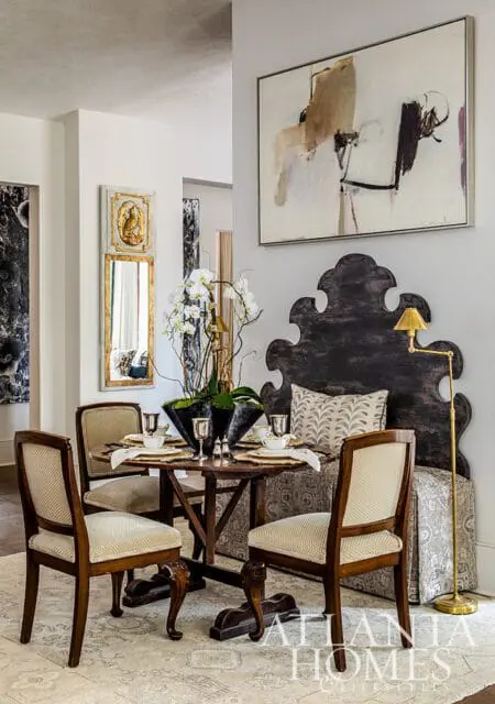 Dining room with a round wooden table set for four, upholstered chairs, a large decorative headboard, and abstract art on the wall. Atlanta Homes & Lifestyles logo in the corner.