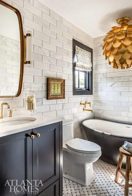 Bathroom with white subway tile walls, dark vanity, white toilet, black clawfoot tub, gold fixtures, and a gold sunburst chandelier.