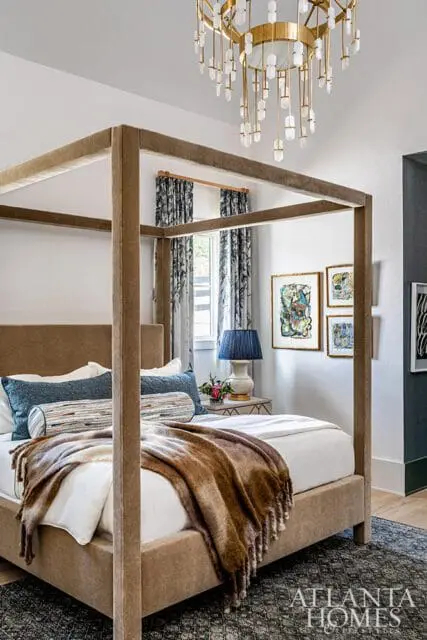 Bedroom with a taupe upholstered four-poster bed, patterned drapes, abstract art, and a gold chandelier.