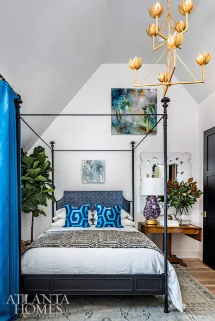 Bedroom with a dark metal four-poster bed, blue patterned headboard and pillows, abstract art, and a gold floral chandelier.