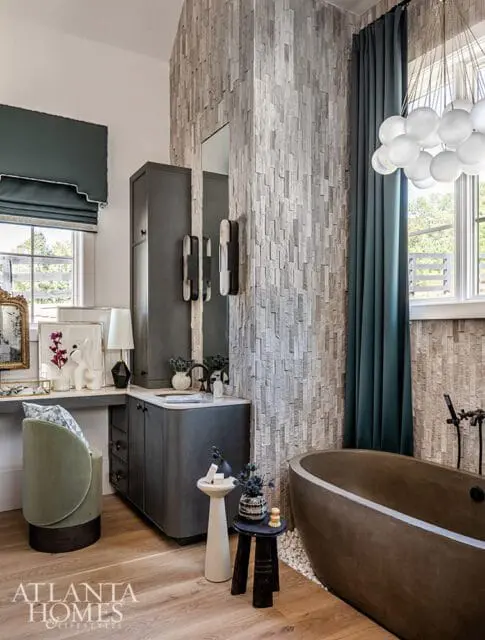 Modern bathroom with a textured stone accent wall, dark gray vanity, freestanding tub, and a green velvet chair.