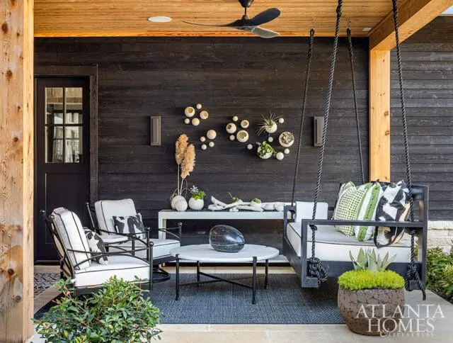 Covered porch with black wood siding, wooden ceiling, and a black swing bed with green and white patterned pillows.