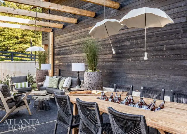Covered outdoor patio with dark wood siding, a long wooden dining table filled with ice and bottled drinks, and seating areas with gray upholstered furniture.