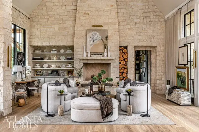 Living room with stone walls, a large stone fireplace with a herringbone brick hearth, built-in shelves, and light gray upholstered furniture including curved sofas and an ottoman.