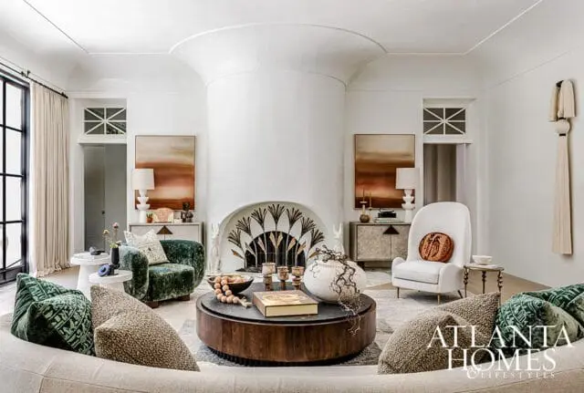 Living room with a large white fireplace featuring a decorative fan motif, two abstract landscape paintings, plush green and cream armchairs, and a dark wood coffee table with decorative objects.
