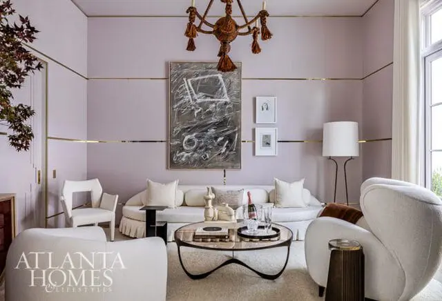 Living room with pale lavender walls, gold trim, white curved sofas, abstract art, and a rope chandelier.