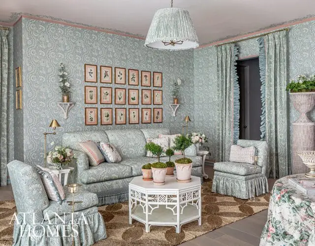 Light blue floral wallpaper adorns this living room, featuring a sofa and chairs upholstered in matching fabric. A collection of botanical prints hangs on the wall above the sofa, and a white hexagonal coffee table sits on a patterned rug.