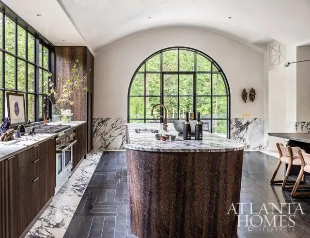 Modern kitchen with a curved island topped with marble, dark wood cabinetry, marble countertops, and a large arched window overlooking a forest.