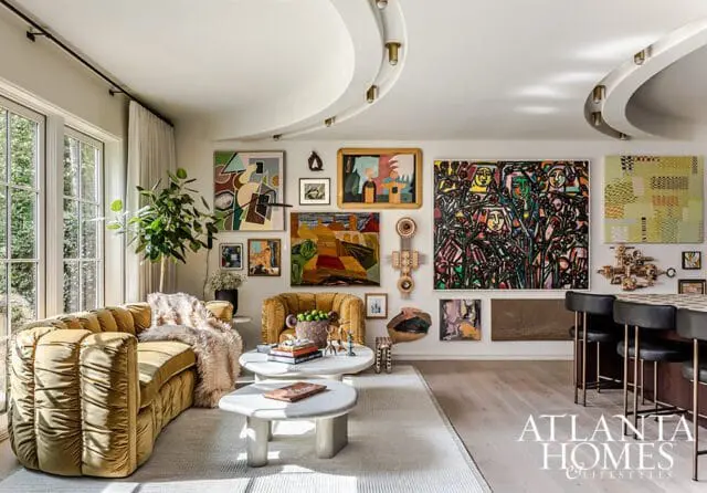 Living room with mustard yellow tufted sofa, abstract art collection on walls, and a curved ceiling with track lighting. A bar area with black stools is visible on the right.