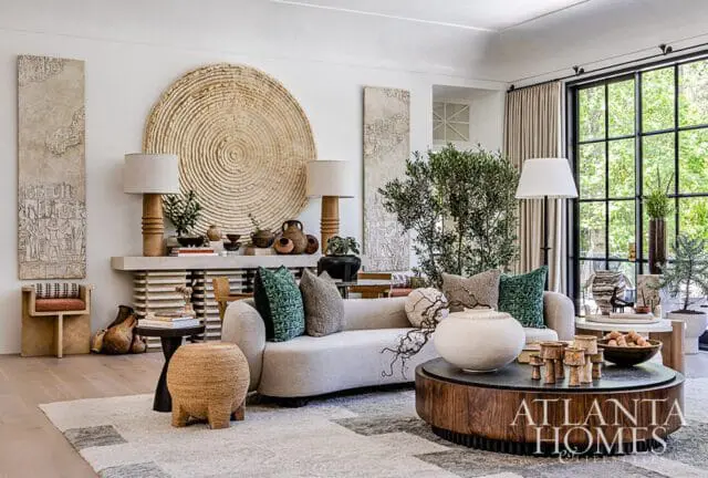 Living room with a curved light gray sofa, textured pillows, a large round woven wall hanging, and a wooden coffee table with decorative objects. A large window with black mullions overlooks greenery.