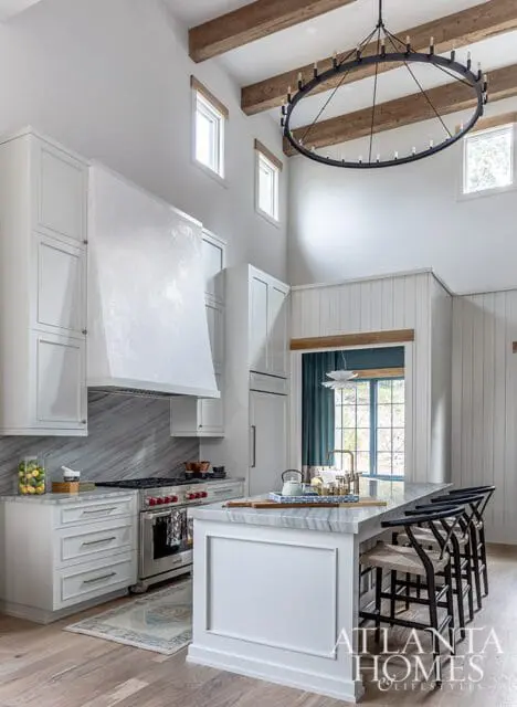 White kitchen with vaulted ceiling, exposed wooden beams, and a large circular chandelier. Features white cabinetry, marble backsplash, and a large island with seating.