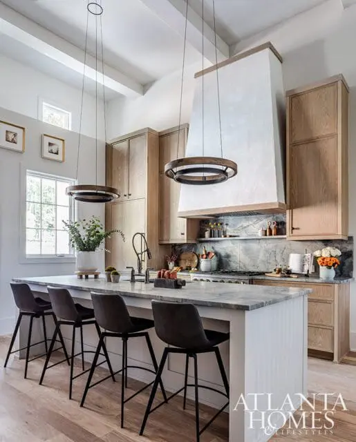Modern kitchen with light wood cabinetry, marble island and countertops, a large plaster range hood, and circular pendant lights.