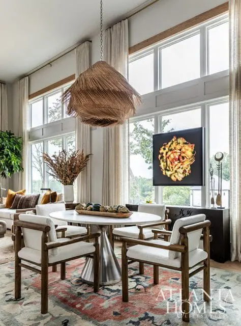 Dining area with a round white marble table, four upholstered chairs with wooden frames, and a large woven pendant light. A colorful rug is on the floor, and large windows overlook greenery.