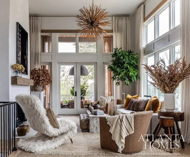 Living room with high ceilings, large windows, a starburst chandelier, a white shag chair, brown armchairs, and a sofa with rust-colored pillows.