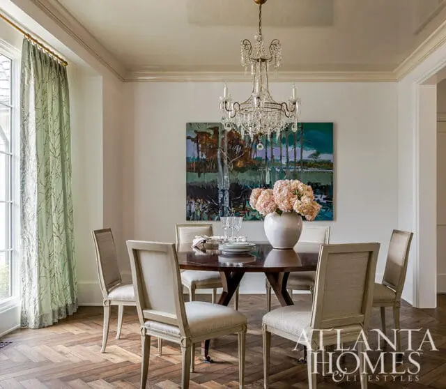 Dining room with a round wooden table set for six, upholstered chairs, a crystal chandelier, and a large abstract painting above the table.