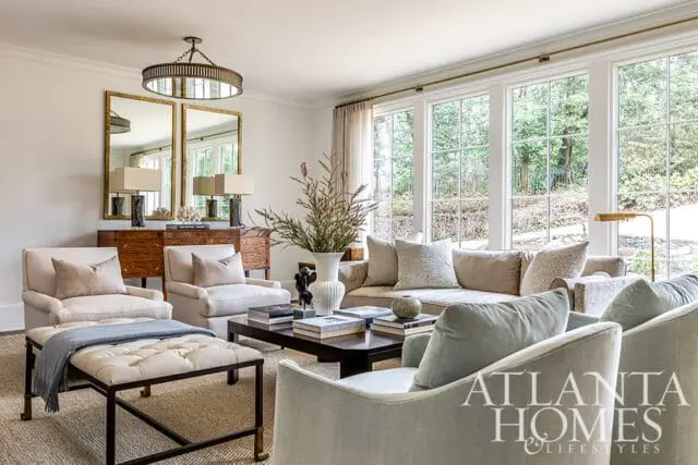 Living room with neutral-toned sofas and armchairs, a dark wood coffee table, and a mirrored cabinet with lamps. Large windows overlook a wooded area.