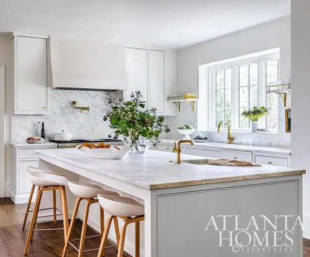 White kitchen with marble countertops and backsplash, brass fixtures, white cabinetry, and a large island with white stools.