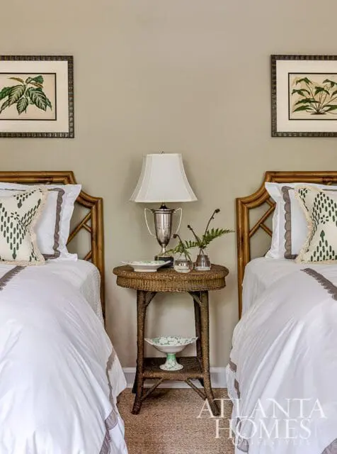Bedroom with two twin beds featuring bamboo headboards, white bedding with grey trim, and botanical prints on the wall. A wicker side table holds a lamp, trophy, and vases.