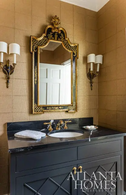 Powder room with gold grasscloth wallpaper, black vanity with a marble countertop, brass fixtures, and an ornate black and gold mirror.