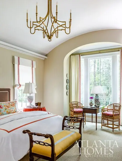 Bedroom with a gold chandelier, white bedding with orange trim, a yellow bench, and rattan chairs by a window overlooking trees.