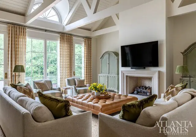 Living room with vaulted ceiling, exposed beams, large windows with patterned curtains, a fireplace with a TV above, a large tufted leather ottoman, and multiple sofas and armchairs.
