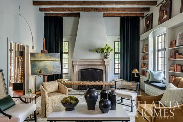 Living room with stone fireplace, dark green curtains, exposed wooden beams, and upholstered furniture. A painting rests on an easel, and built-in bookshelves line the wall.