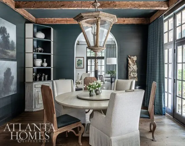 Dark teal dining room with a round light wood table, upholstered chairs, a tall distressed cabinet, and a large ornate lantern hanging from wood beams.
