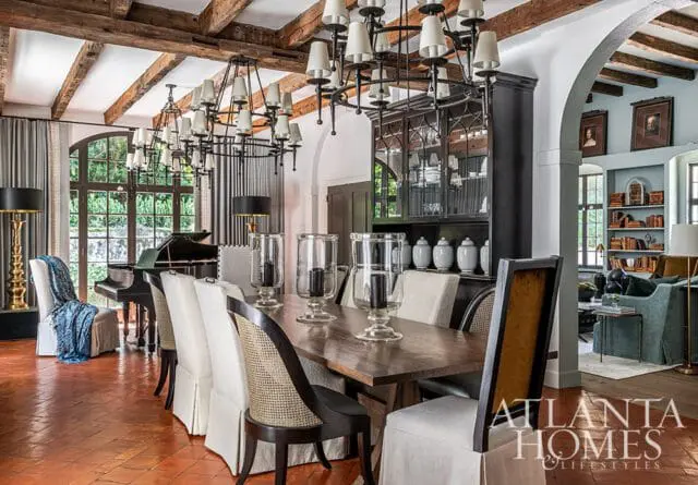 Dining room with a long wooden table, upholstered chairs, and a grand piano near a large window. Exposed wooden beams adorn the ceiling, and a dark china cabinet stands against the wall.