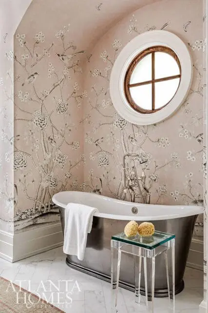 Bathroom with pink floral wallpaper, a freestanding metallic bathtub, and a round window with wooden trim.