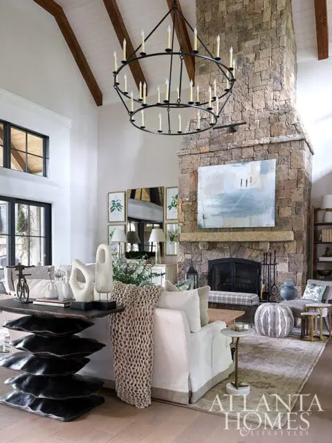 Living room with vaulted ceiling, exposed wooden beams, and a large stone fireplace. A circular chandelier hangs above a sectional sofa and a dark wood console table with sculptural decor.