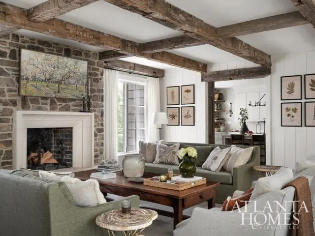 Living room with stone fireplace, exposed wooden beams, green sofas, and botanical prints on the wall.