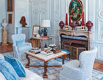 Light blue ornate living room with upholstered armchairs, a patterned rug, a marble fireplace, and decorative urns.