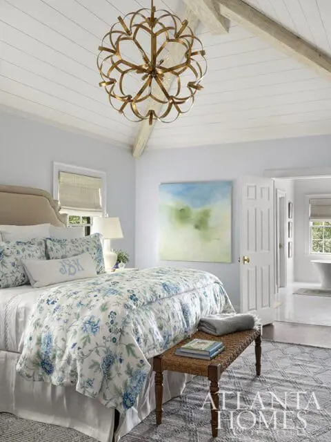 Bedroom with vaulted ceiling, exposed beams, and a large gold chandelier. Bed is dressed in blue floral linens, with a woven bench at the foot. An abstract landscape painting hangs on the wall.