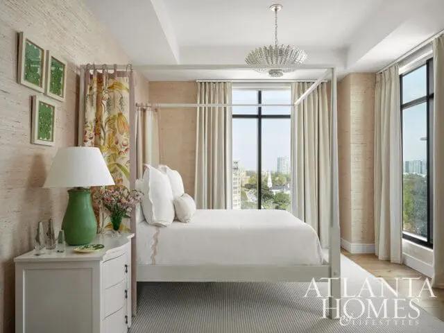 Bedroom with a white four-poster bed, textured beige walls, and large windows overlooking a city skyline. A green lamp sits on a white nightstand.