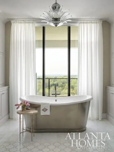 Freestanding bathtub in a neutral-toned bathroom with white curtains, a glass chandelier, and a view of trees through the window.