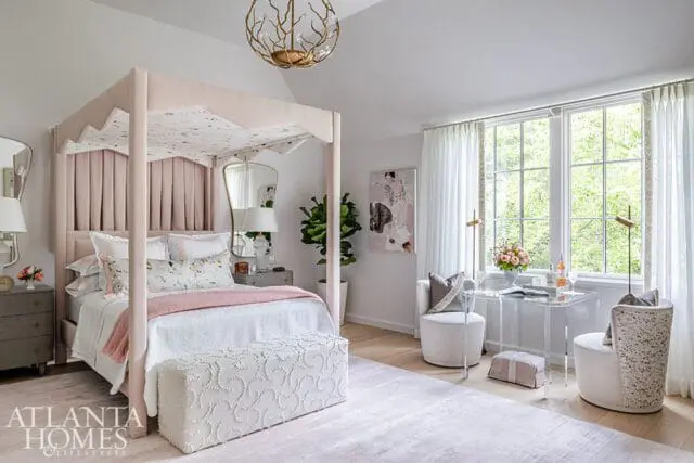 Pink upholstered canopy bed with patterned bedding in a light-filled bedroom. A clear acrylic desk with chairs sits by the window, overlooking lush greenery.