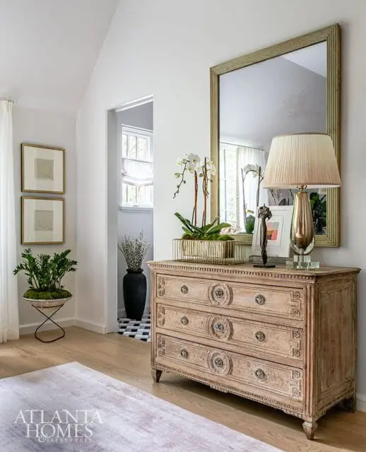 Light wood dresser with ornate carvings and three drawers, topped with orchids, a lamp, and decorative objects. A large framed mirror hangs above.