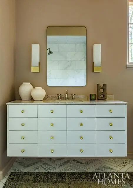 Bathroom vanity with white drawers and gold hardware, marble countertop, and a large rectangular mirror reflecting a marble tiled shower.