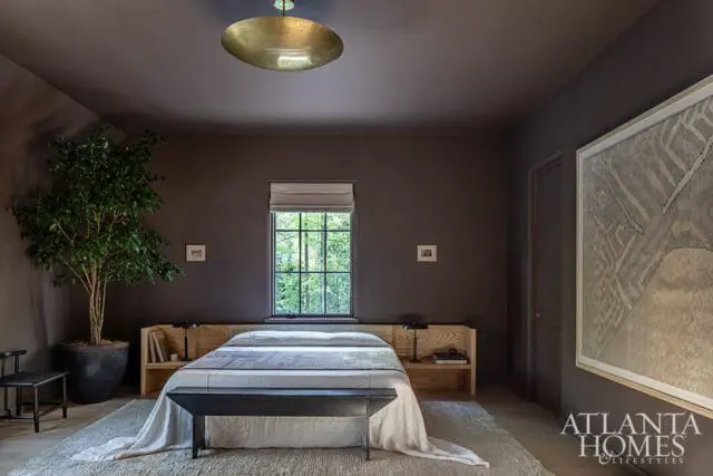 Dark gray bedroom with a queen-size bed featuring a light wood headboard with built-in shelves, a white duvet, and a dark bench at the foot. A large potted tree sits to the left, and a framed abstract artwork hangs on the right wall.