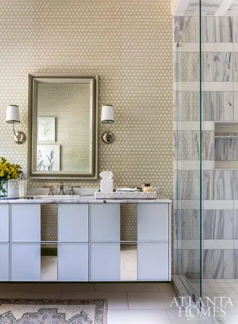 Light blue vanity with mirrored panels below a framed mirror with wall sconces, patterned wallpaper, and marble shower tiles.