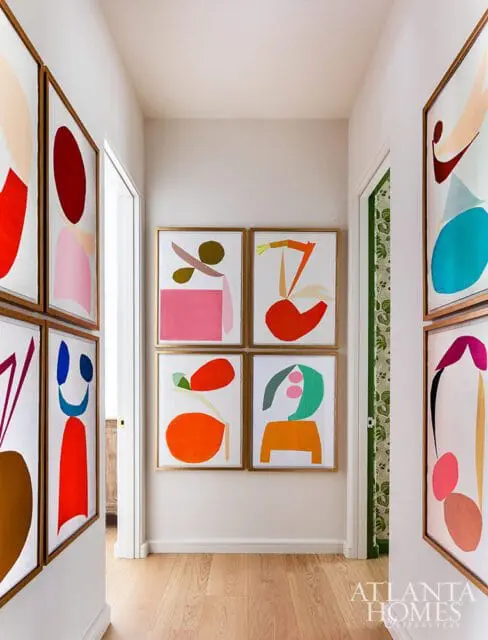 Hallway with light wood floors and walls adorned with framed abstract art featuring bold colors and shapes. A doorway with patterned wallpaper is visible.