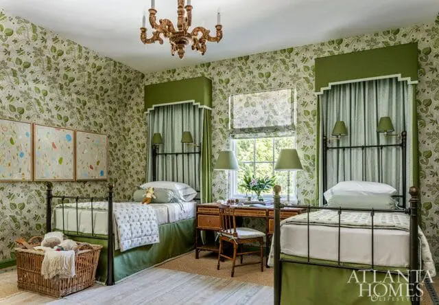 Children's bedroom with green floral wallpaper, two twin beds with green bed skirts, a wooden desk with lamps, and a brass chandelier.