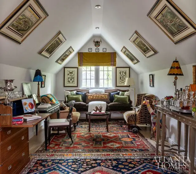 Attic living room with vaulted ceiling, featuring a dark brown sofa with colorful pillows, a large Persian rug, a wooden desk, and framed artwork on the walls and ceiling.