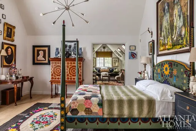 Bedroom with a four-poster bed featuring a colorful, patterned headboard and a patchwork quilt. Artwork adorns the walls, and a doorway reveals a glimpse of a living area.