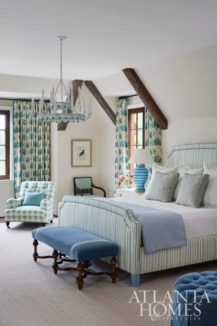 Bedroom with a striped bed, blue upholstered bench, plaid armchair, and a chandelier. The room features blue and white floral curtains and exposed wooden beams.