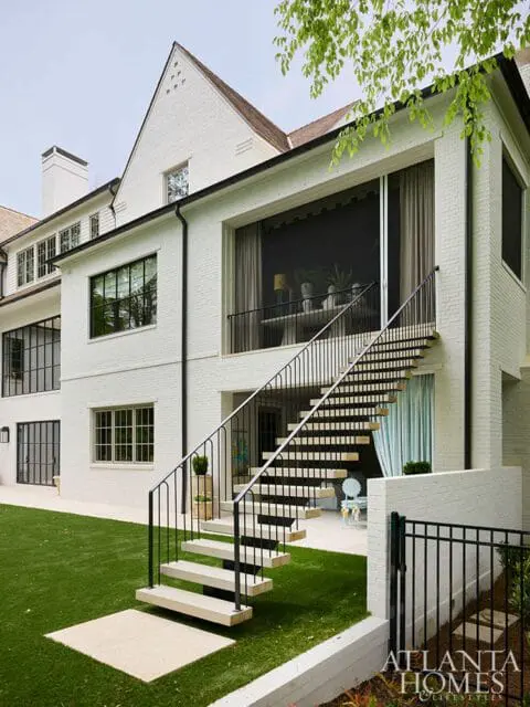 White brick house with black framed windows and a modern floating staircase leading to a screened-in porch. Artificial turf lawn in foreground.