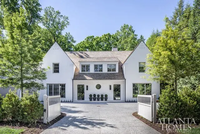 White stucco house with gabled roofs, dark-framed windows, and a circular detail above the entrance. A gravel driveway leads to the house, flanked by white gates and mature trees.