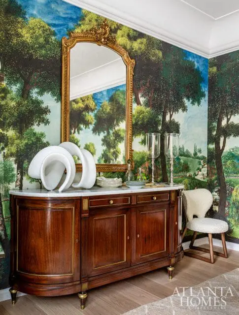 Dark wood buffet with marble top, ornate gold mirror, and pastoral wallpaper. A sheepskin chair sits to the right.
