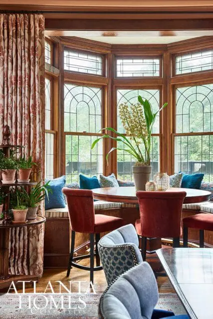 Dining nook with built-in banquette seating, round wooden table, red velvet chairs, and large bay windows with arched panes.