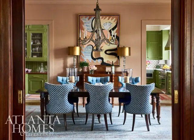 Dining room with dark wood table and chairs upholstered in blue and white patterned fabric, facing a buffet with lamps and abstract art. Green cabinets visible in adjacent kitchen.